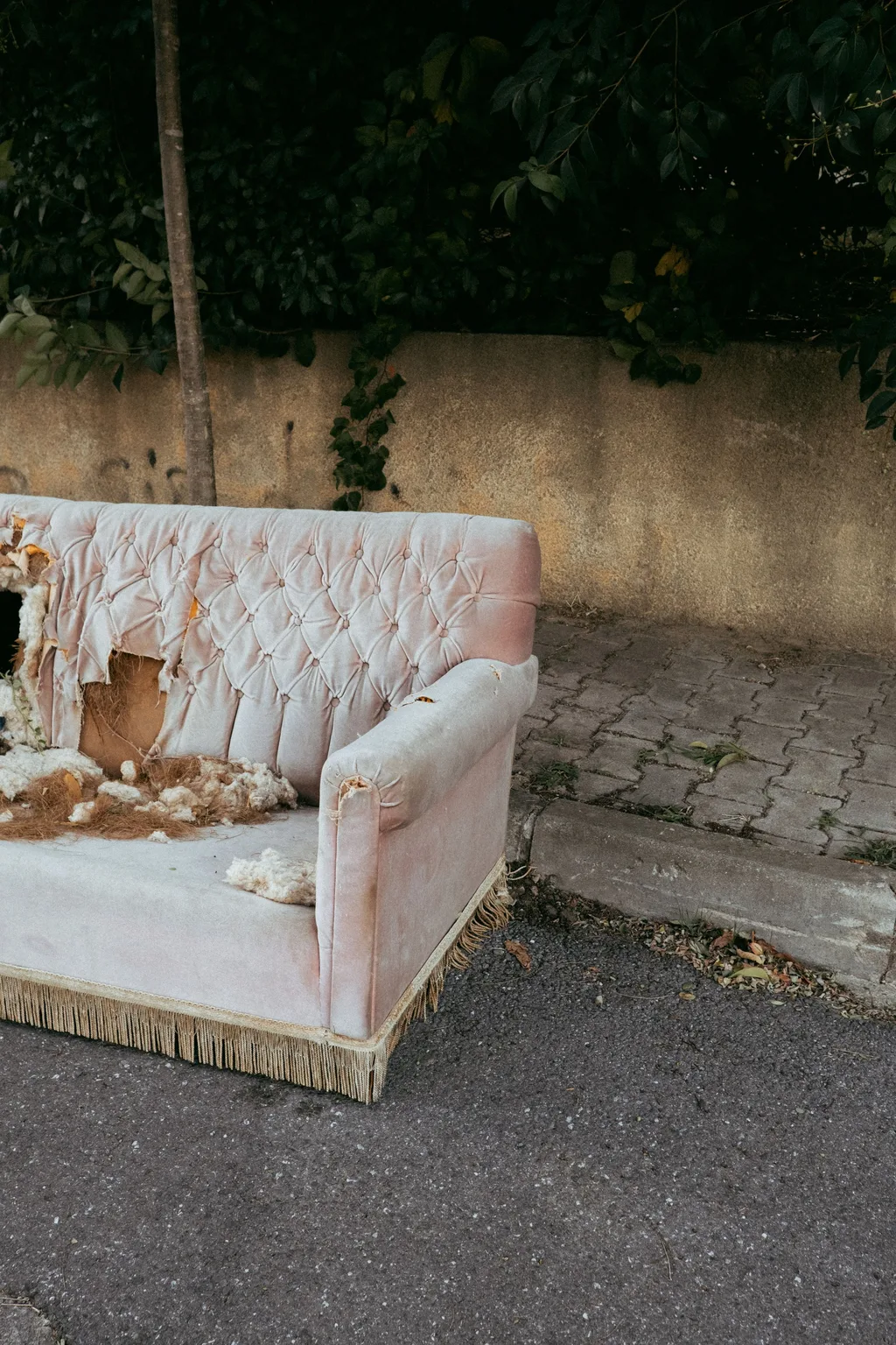 A worn pink buttoned sofa with torn upholstery left at the kerb beside a weathered concrete wall