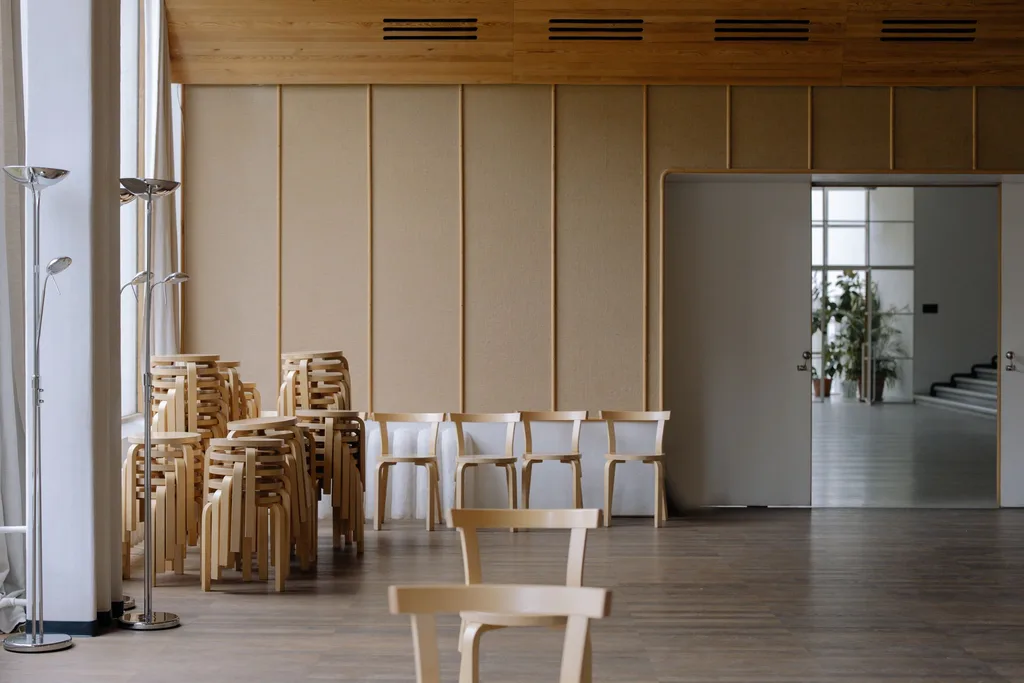 A cleared commercial hall with wooden chairs and stools stacked against one wall beside an open doorway