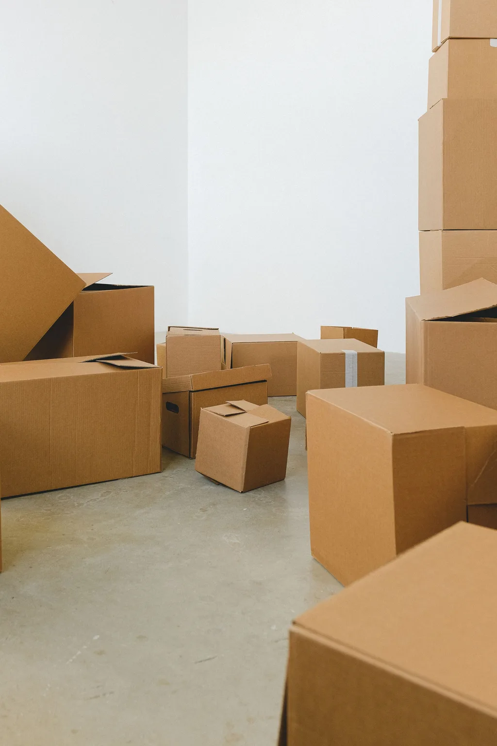 Open and sealed cardboard moving boxes grouped on the bare concrete floor of an otherwise empty white room
