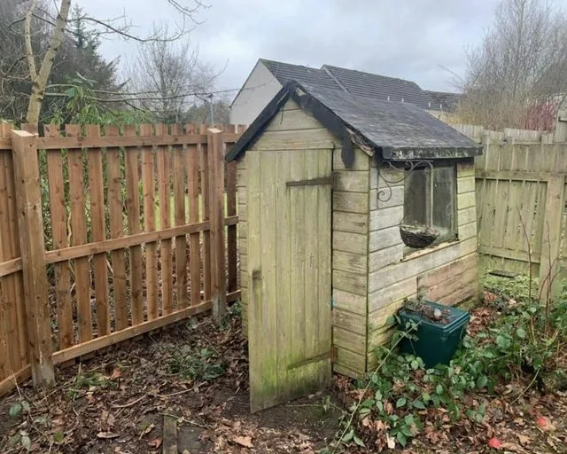 A weathered wooden garden shed with a broken window, standing beside a fence in a back garden